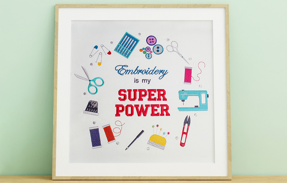 Square wooden frame displaying embroidered fabric with the phrase 'Embroidery is my Super Power' surrounded by various craft-related embroidered designs.