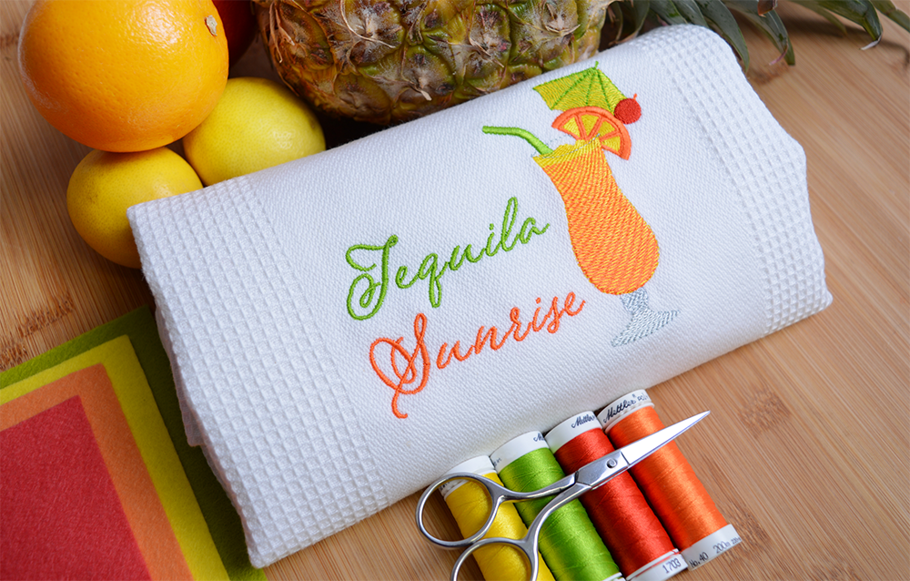 White dishcloth embroidered with 'Tequila Sunrise' lettering and a color-blended cocktail glass design.